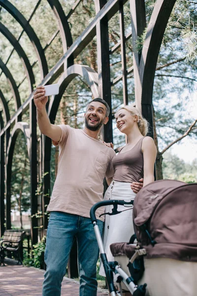 Padres sonrientes tomando selfie con teléfono inteligente cerca de carro de bebé en el parque - foto de stock