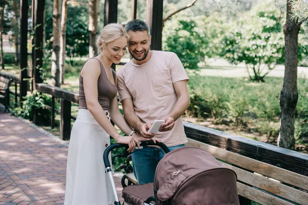 Parents taking photo of baby with smartphone near baby carriage in park — Stock Photo