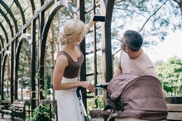 Vue latérale des parents prenant selfie avec smartphone près de la poussette dans le parc — Photo de stock