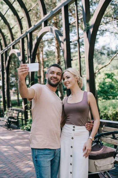 Genitori sorridenti che scattano selfie con smartphone vicino alla carrozzina nel parco — Foto stock