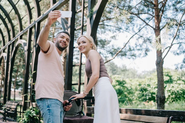 Low angle view of parents taking selfie with smartphone near baby carriage in park — Stock Photo