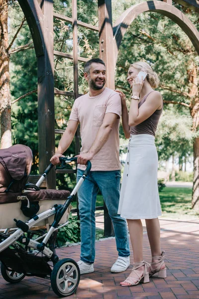 Glückliche Mutter spricht mit Smartphone in der Nähe von Kinderwagen und palmt Ehemann im Park — Stockfoto