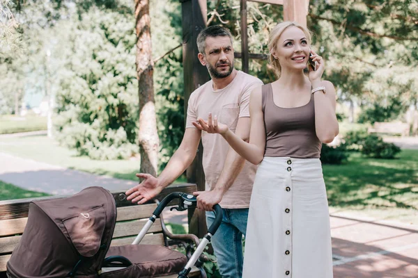 Happy mother talking by smartphone near baby carriage in park, irritated father looking at her — Stock Photo