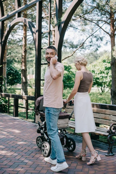 Father talking by smartphone near baby carriage in park — Stock Photo