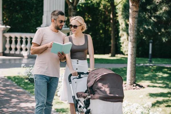 Parents marchant avec la voiture de bébé dans le parc et le livre de lecture — Photo de stock