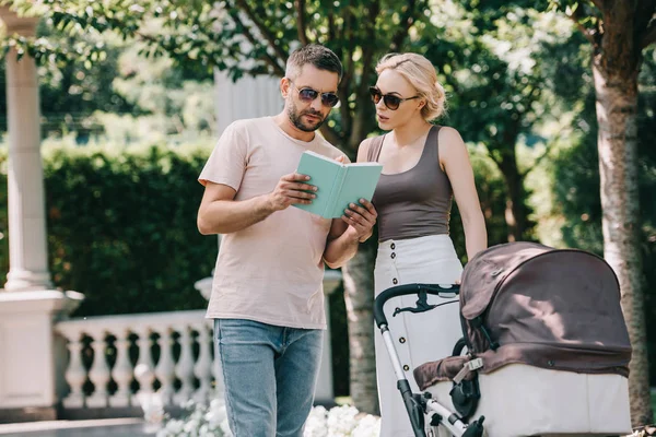 Parents standing near baby carriage in park and reading book — Stock Photo