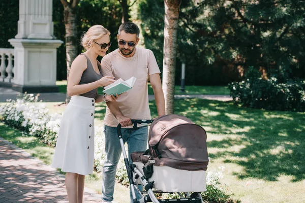 Mother and father standing near baby carriage in park and holding book — Stock Photo