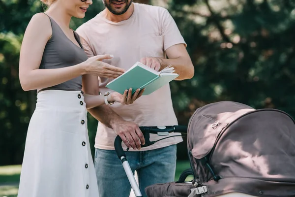Cropped image of parents standing near baby carriage in park and reading book — Stock Photo