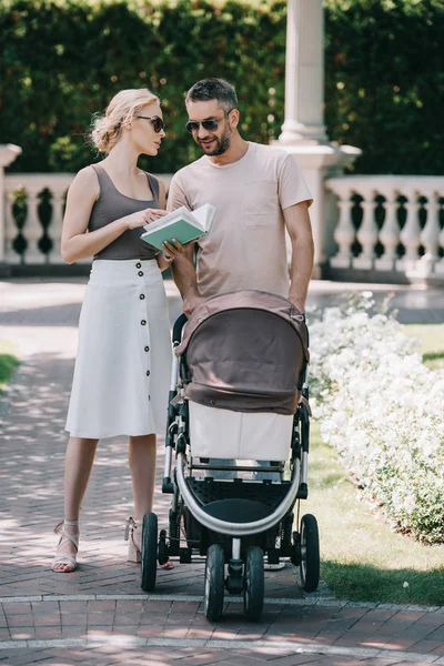 Parents standing near baby carriage in park and holding book — Stock Photo