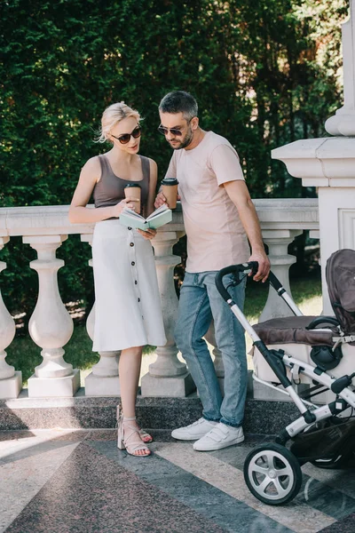 Parents debout près de voiture de bébé dans le parc, livre de lecture et tenant des tasses à café jetables — Photo de stock