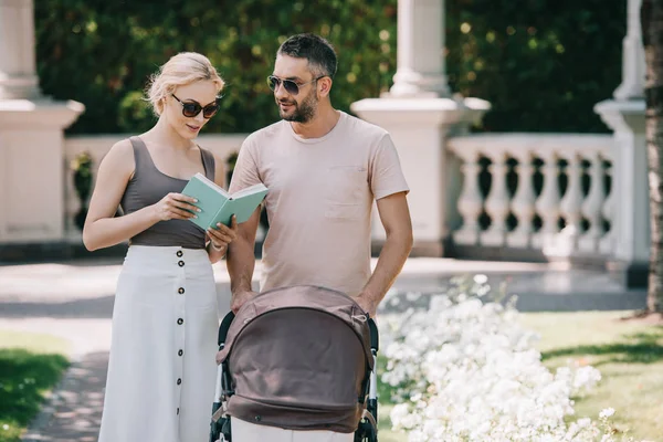 Parents in sunglasses standing near baby carriage in park and reading book — Stock Photo