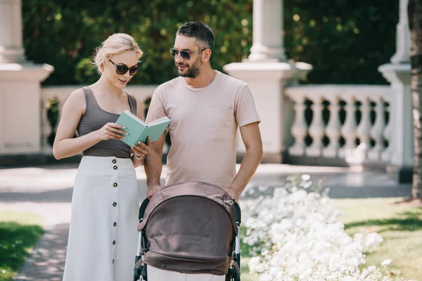 Padres en gafas de sol de pie cerca de carro de bebé en el parque y libro de lectura - foto de stock