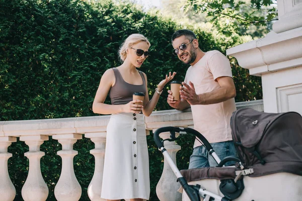Parents holding disposable coffee cups and quarreling near baby carriage in park — Stock Photo