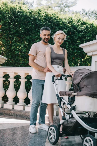 Husband hugging wife near with baby carriage in park and they looking at kid — Stock Photo