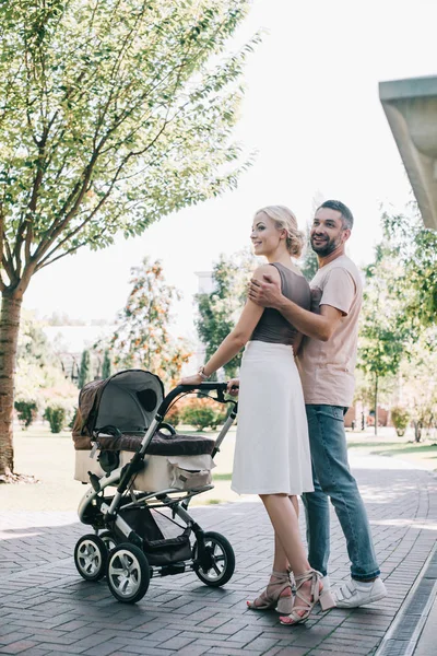 Happy parents cuddling and walking with baby carriage in park — Stock Photo