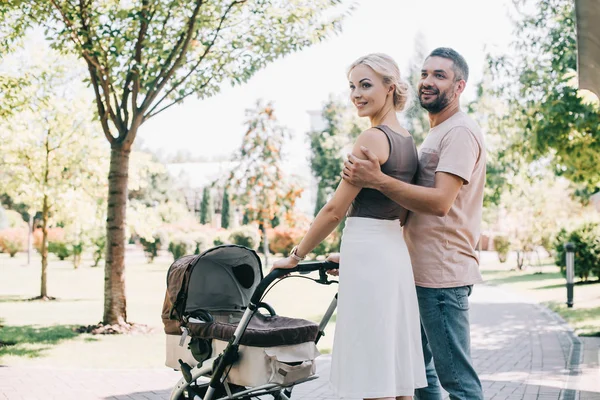 Parents heureux étreignant et marchant avec la voiture de bébé dans le parc — Photo de stock