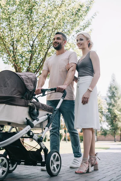 Visão de baixo ângulo de pais felizes andando com carruagem de bebê no parque — Fotografia de Stock