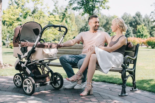Happy parents sitting on bench near baby carriage in park and looking at each other — Stock Photo