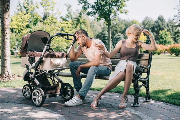 Padres agresivos sentados en el banco cerca del carro del bebé en el parque - foto de stock