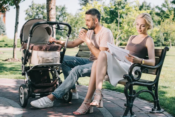 Père buvant du café et touchant bébé chariot dans le parc, mère lisant le journal — Photo de stock