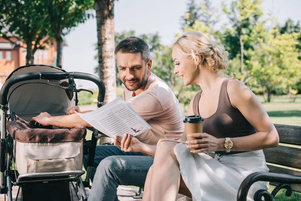 Esposa mostrando periódico a marido en banco cerca de carro de bebé en parque - foto de stock