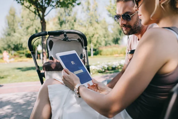 Pais sentados perto de carruagem do bebê no parque e olhando para tablet com página Facebook carregada — Fotografia de Stock