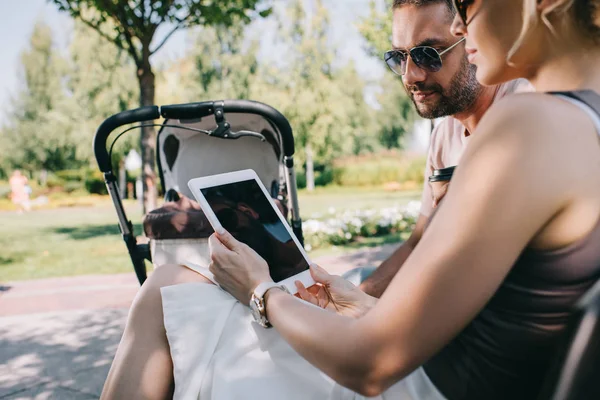 Mother and father sitting near baby carriage in park and looking at tablet with blank screen — Stock Photo