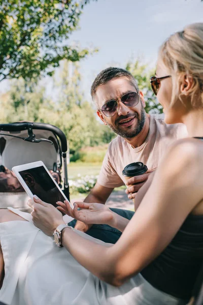 Padres sentados cerca del carro del bebé en el parque y sosteniendo la tableta con pantalla en blanco - foto de stock