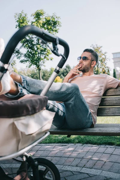 Father smoking cigarette on bench with legs on baby carriage in park — Stock Photo