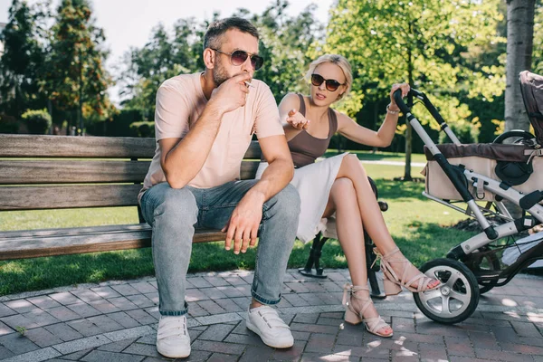 Padre fumando cigarrillo cerca del cochecito de bebé en el parque, madre haciendo gestos y mirándolo - foto de stock