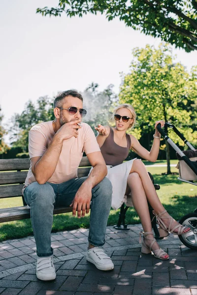 Чоловік палить сигарету біля дитячого перевезення в парку, дружина жестикулює і дивиться на нього — стокове фото