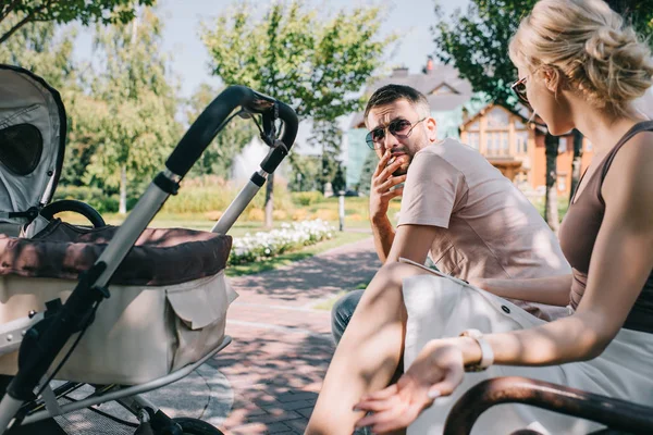 Husband smoking cigarette near baby carriage in park and looking at wife — Stock Photo