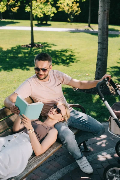 Vista ad alto angolo della moglie sdraiata sulle gambe del marito vicino alla carrozza del bambino nel parco e nel libro di tenuta — Foto stock