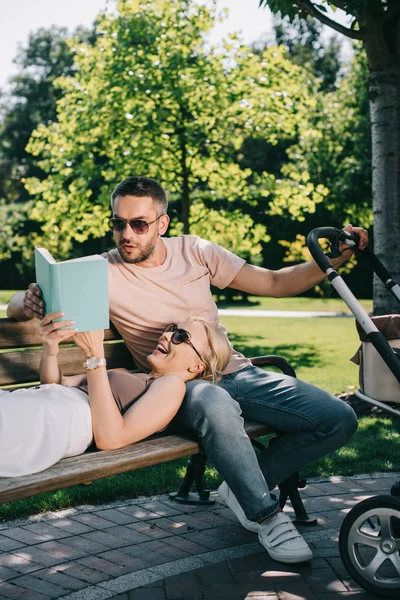 Riendo esposa acostado en marido piernas cerca bebé carro en parque y lectura libro - foto de stock