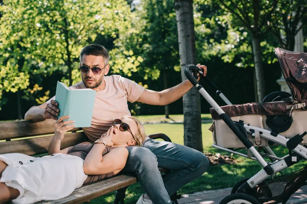 Esposa acostada en las piernas del marido cerca de carro de bebé en el parque y libro de celebración, marido leyendo para ella - foto de stock