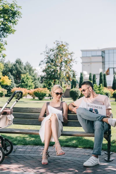 Parents sitting on bench near baby carriage in park and reading magazine and business newspaper — Stock Photo