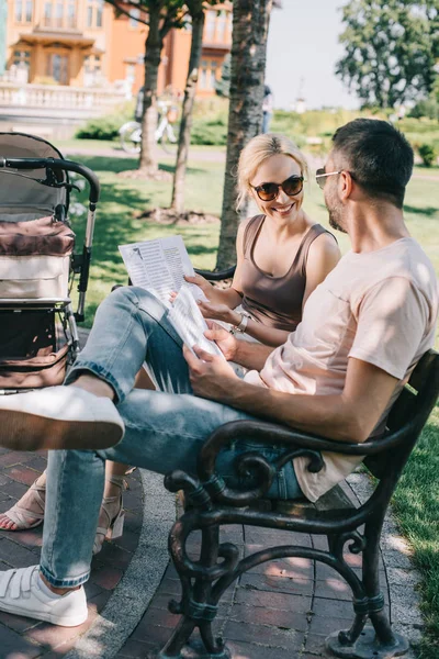 Smiling parents sitting on bench near baby carriage in park with magazine and newspaper — Stock Photo