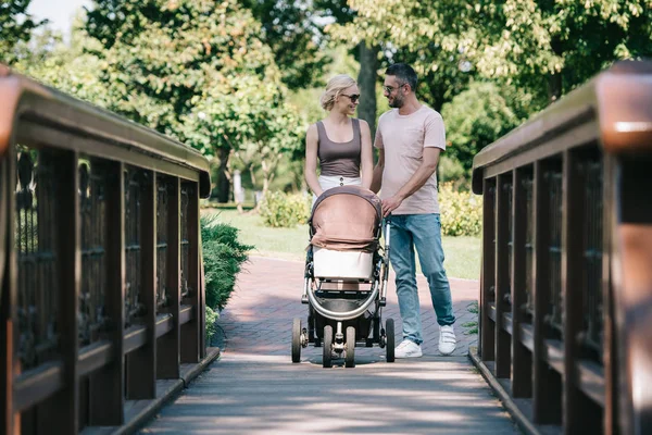 Happy parents walking with baby carriage on bridge in park and looking at each other — Stock Photo