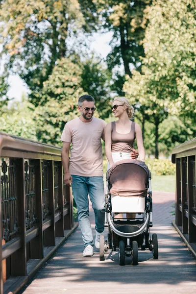 Happy parents walking with baby carriage on bridge in park — Stock Photo