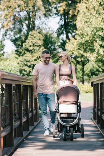 Genitori felici che camminano con carrozzina sul ponte nel parco — Foto stock