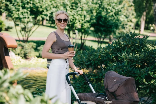 Mère marchant avec la voiture de bébé dans le parc et tenant le café pour aller — Photo de stock