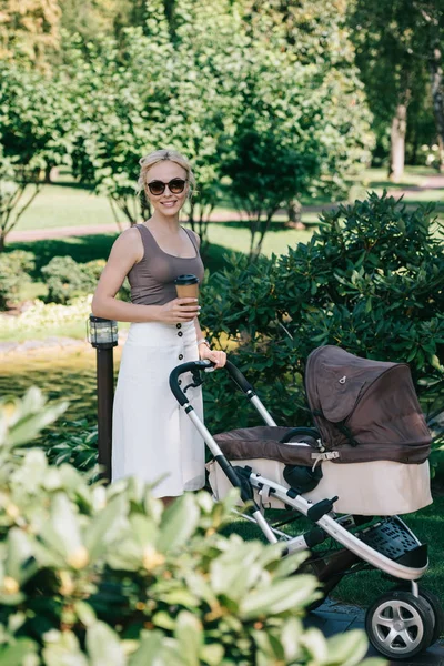 Mère marchant avec la voiture de bébé dans le parc et tenant le café dans la tasse en papier — Photo de stock