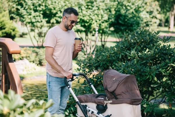 Père marchant avec la voiture de bébé dans le parc et tenant le café dans la tasse en papier — Photo de stock