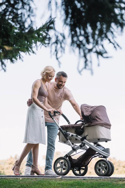 Happy parents walking with baby carriage and looking at baby in park — Stock Photo