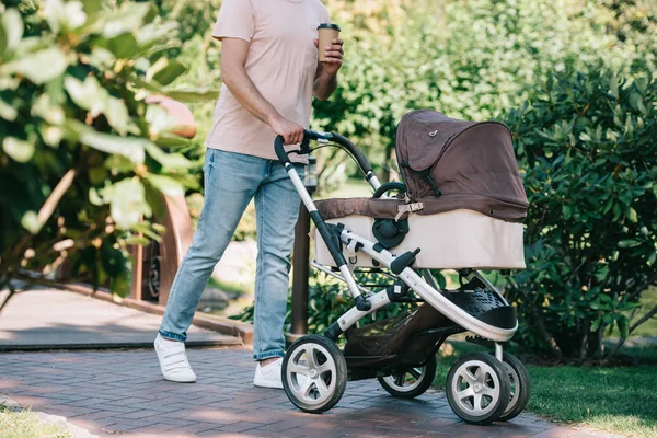 Abgeschnittenes Bild eines Vaters, der mit Kinderwagen und Einweg-Kaffeetasse im Park spaziert — Stockfoto