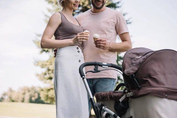 Cropped image of parents holding ice cream and looking at baby carriage in park — Stock Photo