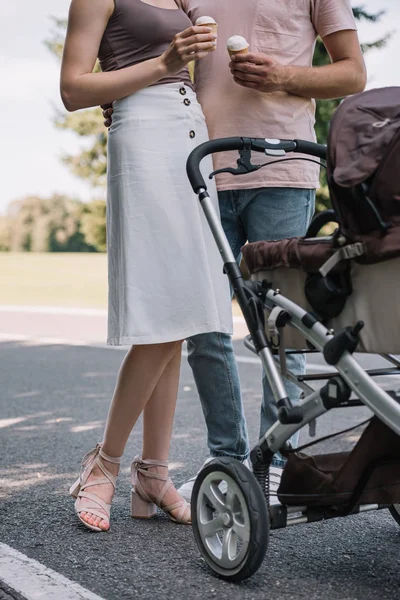 Cropped image of parents holding ice cream and standing near baby carriage in park — Stock Photo