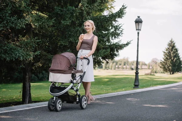 Sonriente madre caminando con carro de bebé y comiendo helado en el parque - foto de stock