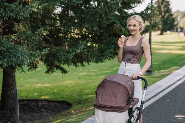 Smiling mother walking with baby carriage and ice cream in park — Stock Photo