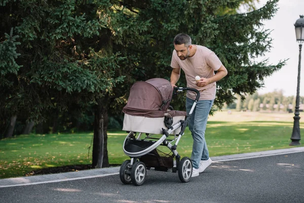 Smiling father walking with baby carriage and ice cream in park and looking at baby — Stock Photo